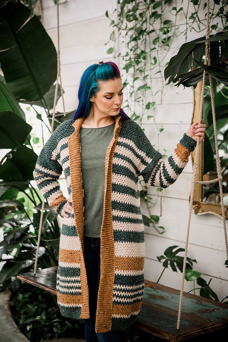 A person with multicolored hair stands on a wooden swing, wearing a striped crocheted cardigan and unique jewellery, surrounded by green, leafy plants.