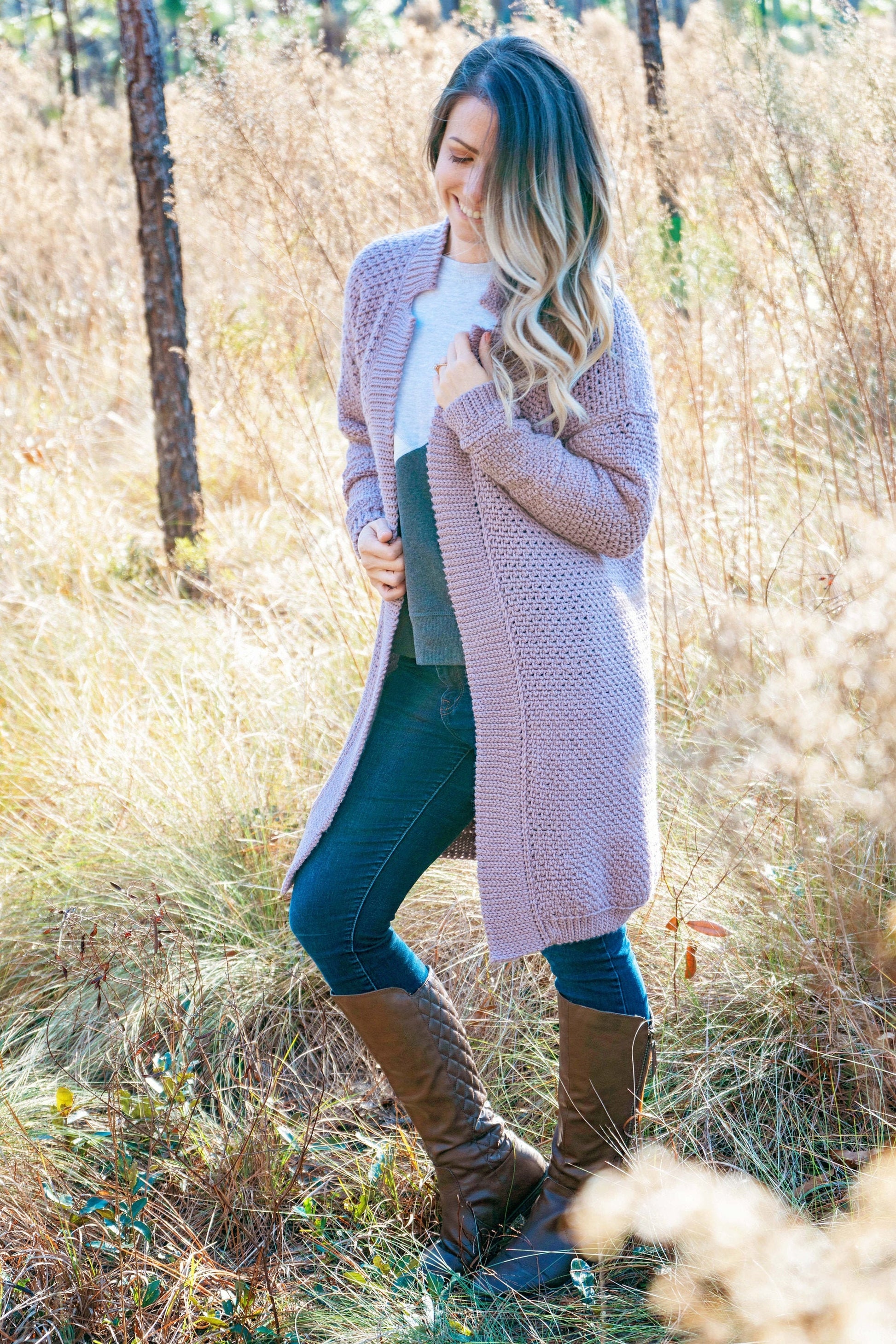 A woman stands in an autumn field wearing a long, knit cardigan, jeans, and tall boots. She looks down with one hand touching her hair, showcasing unique jewellery that complements her vintage clothes.