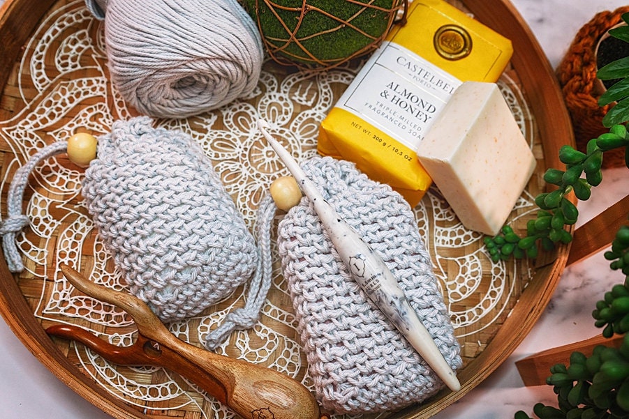 Wooden tray with crochet soap savers, a soap bar, crochet hooks, and ball of yarn on a lace doily. Green plant decor is partially visible on the side. Perfect for those who appreciate unique jewellery or seek a charming handmade gift.
