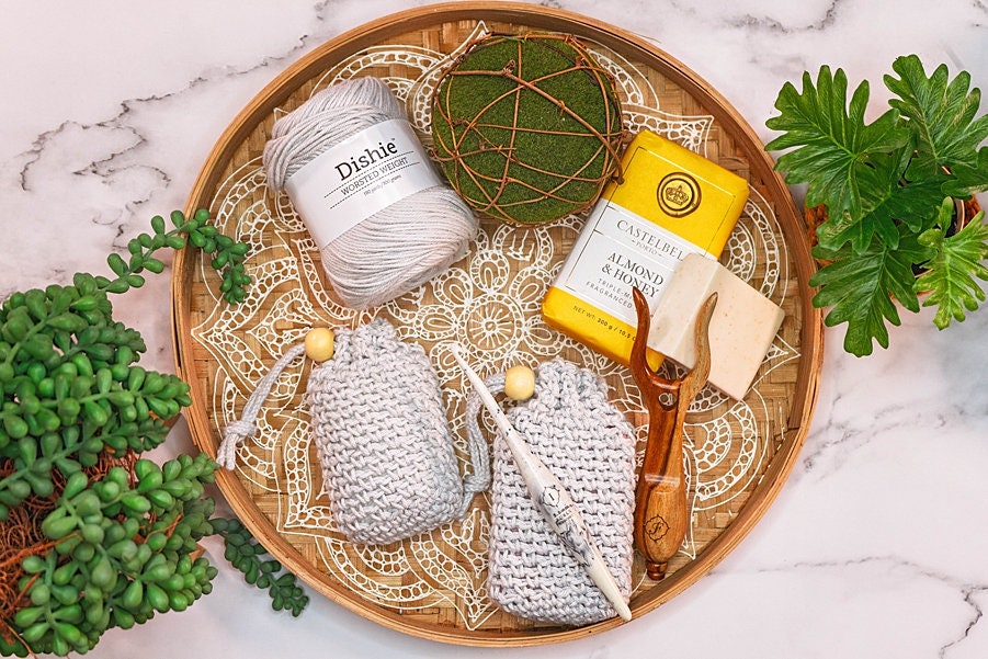 A round wooden tray displays gray yarn, crochet hooks, crocheted soap holders, almond butter soap, and a plant arrangement— a perfect setting for showcasing handmade gifts. Small green plants are placed around the tray.