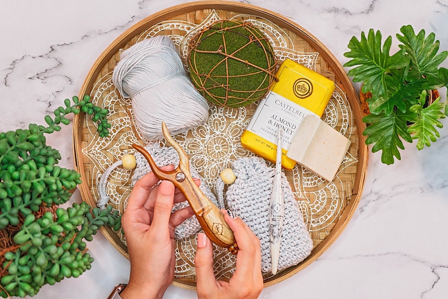Hands holding a crochet hook over a tray with yarn, knitted pieces, a bar of soap, and unique jewellery as a green accessory glints beside two potted plants on the marble surface.