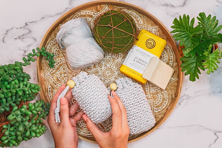 Two hands knitting with white yarn, surrounded by plants, soap, and decorative items—ideal for creating a handmade gift—on a patterned circular tray on a marble surface.
