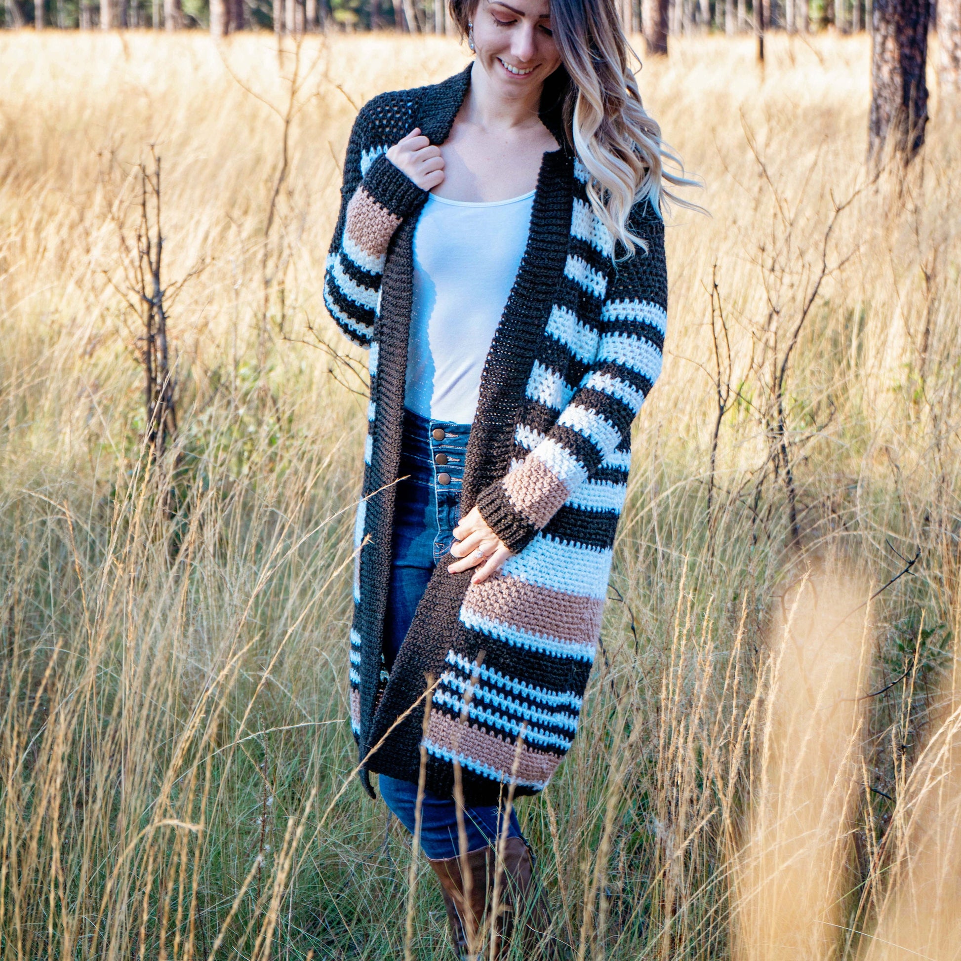 A woman stands in a grassy field, wearing a white top, blue jeans, and a long striped cardigan. She completes her look with unique jewellery, adding a touch of individuality to her ensemble.