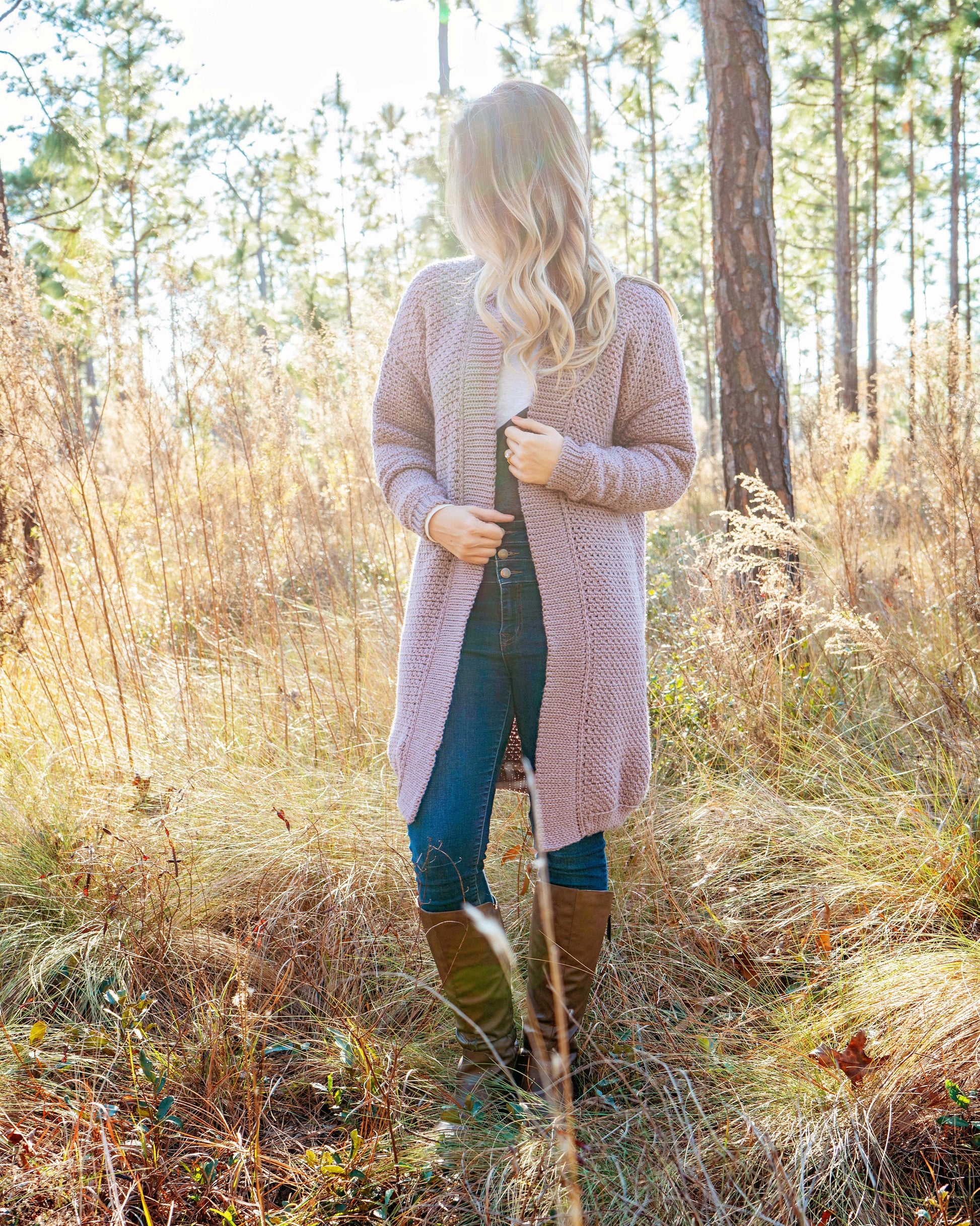 A person standing in a field of tall grass surrounded by trees, wearing a long cardigan, vintage clothes, jeans, and boots, facing slightly away from the camera.