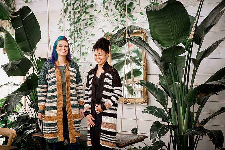 Two people wearing vintage clothes stand in a greenery-filled room with large plants and a mirror on the wall, their outfits complemented by pieces of unique jewellery.