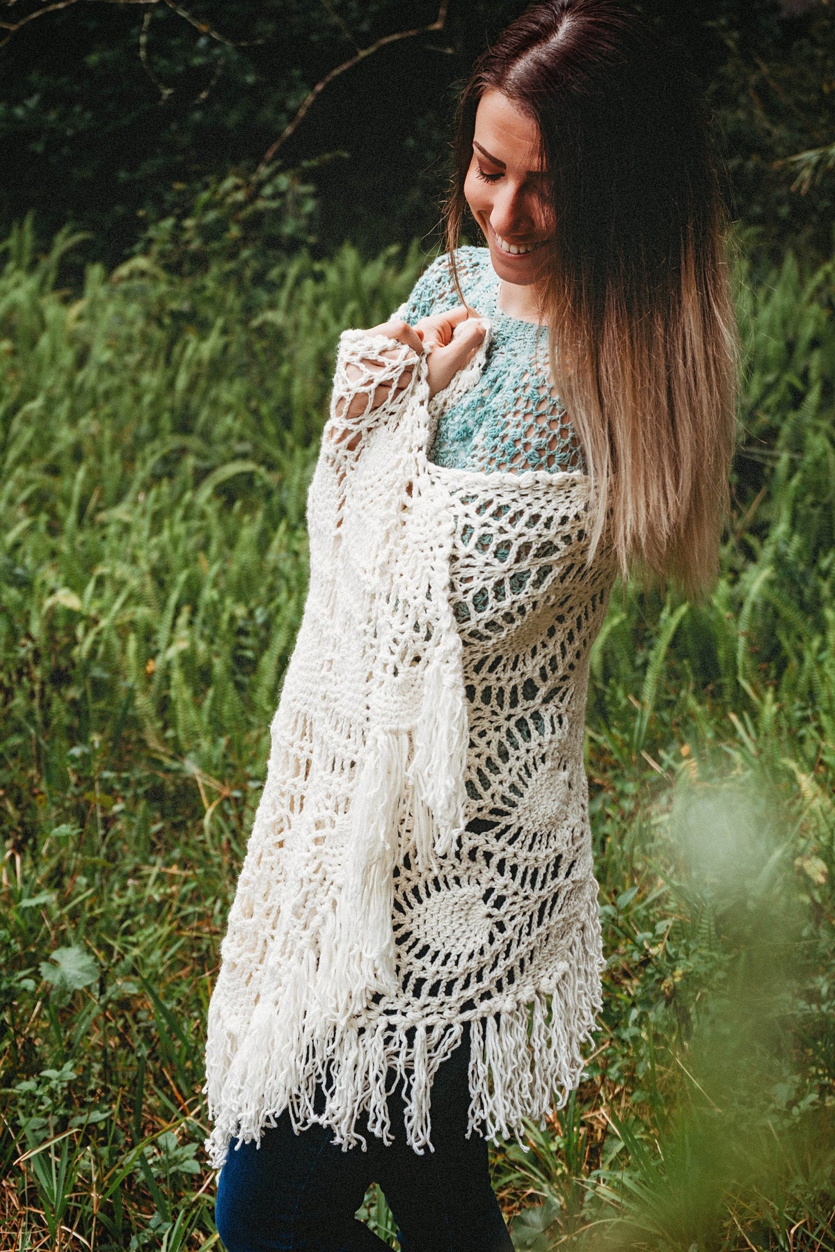 A woman wearing a white shawl decorated with the intricate crochet pineapple stitch, with fringed edges. She stands in an outdoor green space, surrounded by greenery and sunshine.