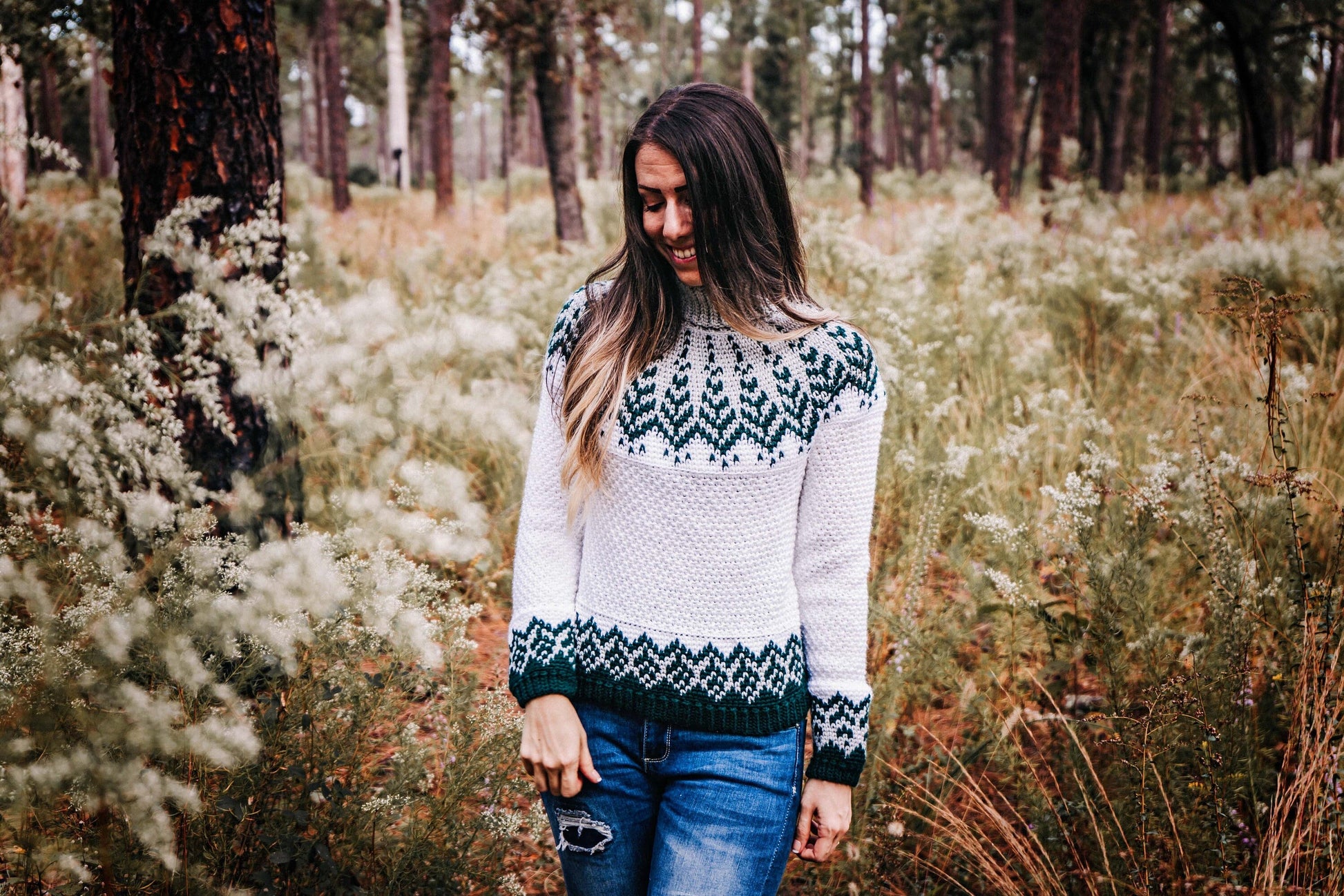 A woman, wearing a patterned sweater and jeans, stands amidst tall grass and wildflowers in a forested area, smiling down as she admires the unique jewellery she received as a handmade gift.