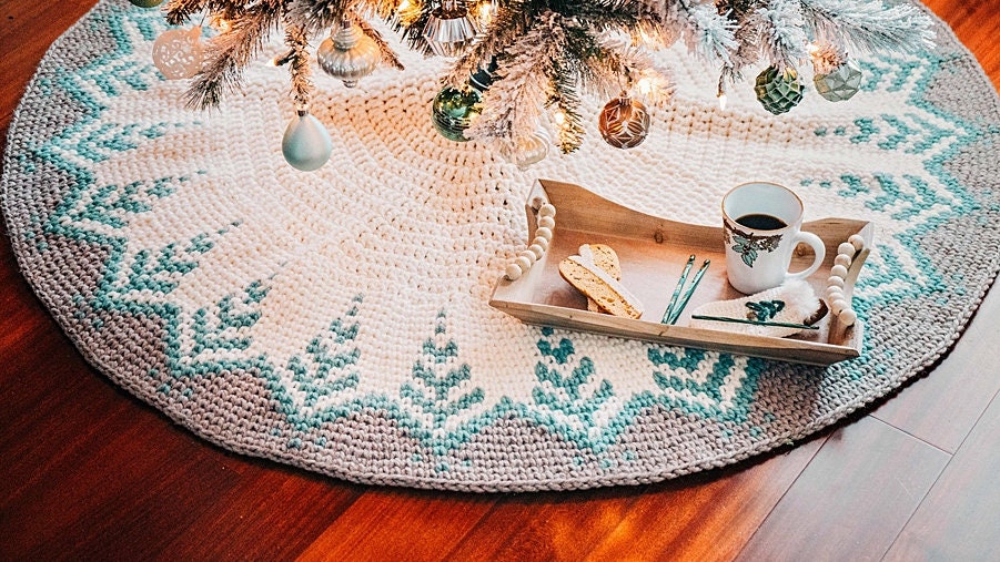 A crocheted tree skirt with a snowflake pattern in white and teal sits under a decorated Christmas tree, resembling vintage clothes. A wooden tray holds a cup of coffee and cookies on the skirt, perfect for enjoying.