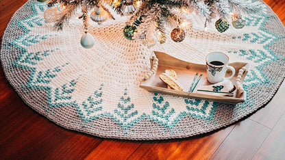 A crocheted tree skirt with a snowflake pattern in white and teal sits under a decorated Christmas tree, resembling vintage clothes. A wooden tray holds a cup of coffee and cookies on the skirt, perfect for enjoying.