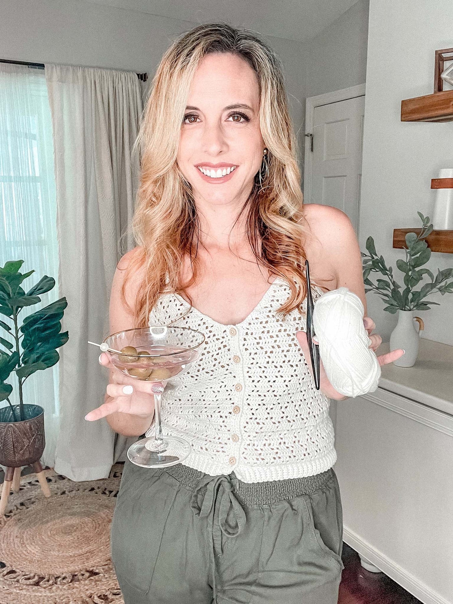 A woman stands indoors, holding a martini glass with olives in one hand and a ball of white yarn in the other. She is smiling and wears vintage clothes, including a sleeveless button-up top and green pants.