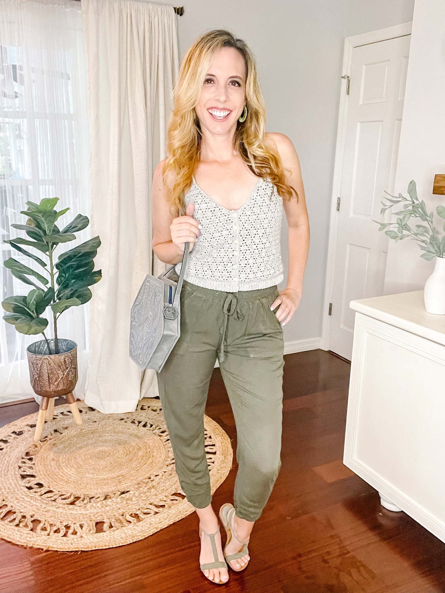 A woman with long blond hair stands in a room, wearing a white sleeveless top and green pants, holding a gray handbag. She is smiling and there are plants, a circular rug in the background.