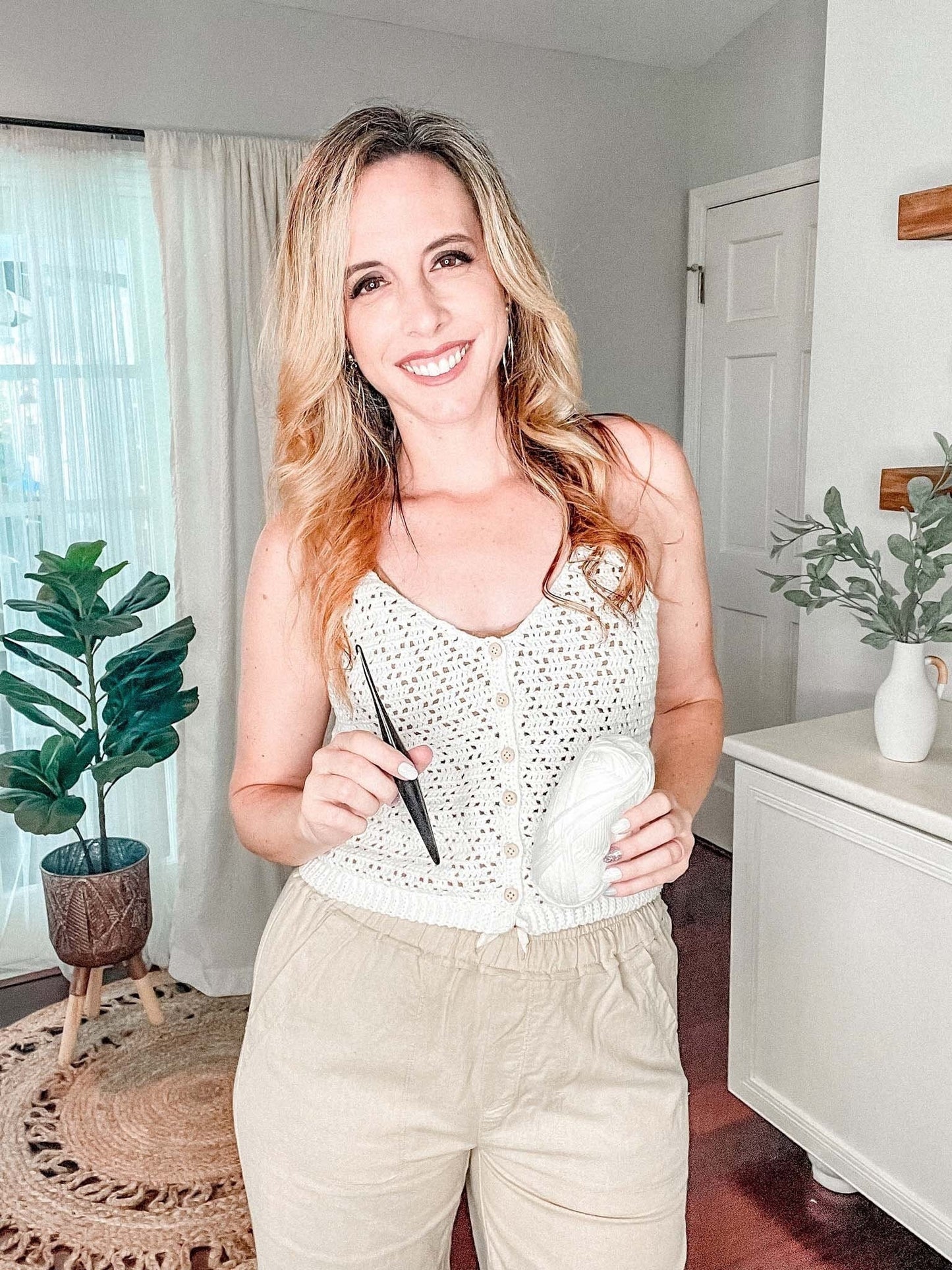A woman with long blonde hair stands indoors smiling, holding a white skein of yarn and a crochet hook, wearing a knitted white top and beige pants. Plants and home decor are visible in the background.