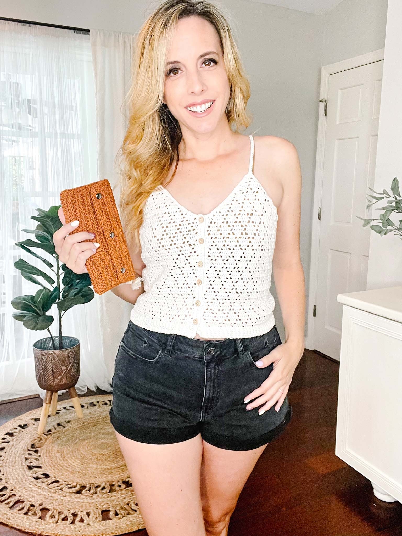 A woman with long blonde hair stands indoors, wearing a white crochet top and black shorts. In her left hand, she holds a brown clutch. A potted plant and white furniture are in the background.