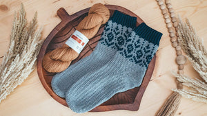 Pair of gray crochet socks with dark teal colorwork cuffs on a wooden leaf tray with a tan yarn skein