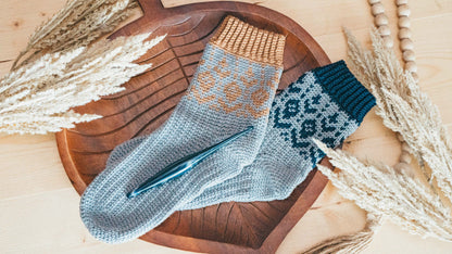Gray crochet socks with tan and teal patterned cuffs and a crochet hook on a wooden tray with dried grass