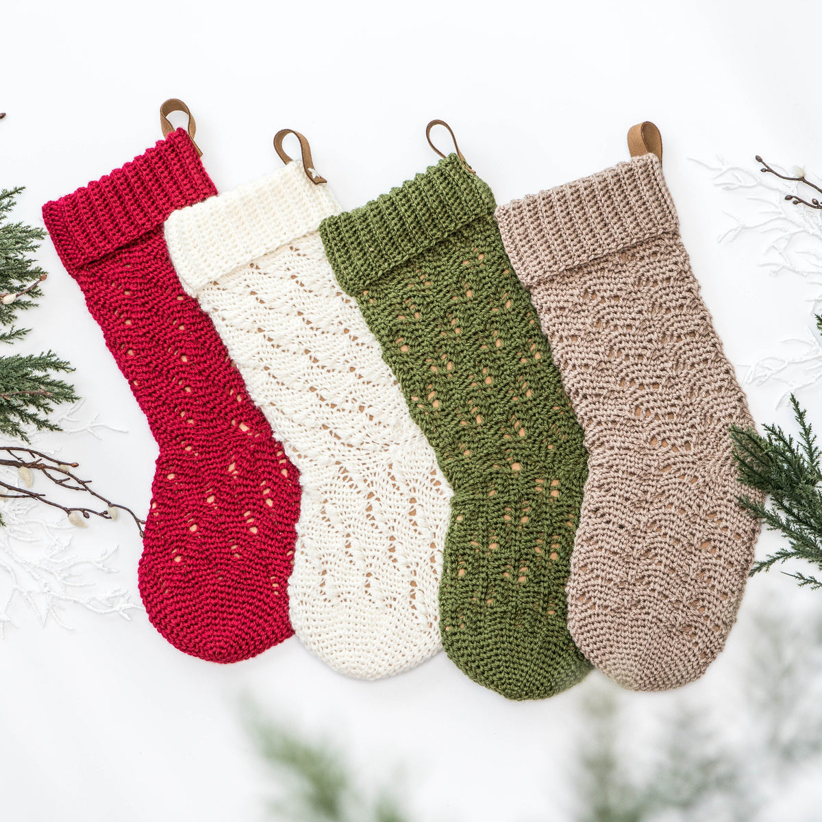 Four crocheted Christmas stockings in red, white, green, and beige hang side by side, surrounded by decorative evergreen branches. These charming pieces make a thoughtful handmade gift that captures the warmth of the season.