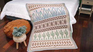 Close view of cream crochet blanket with blue mountain, brown geometric and green cactus rows hanging off a bed