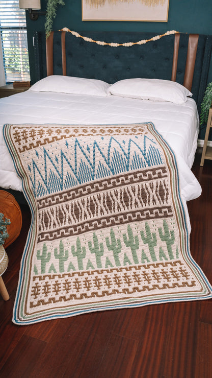 Crochet mosaic blanket in cream, blue, brown and sage green with striped border spread over a white bed onto the floor