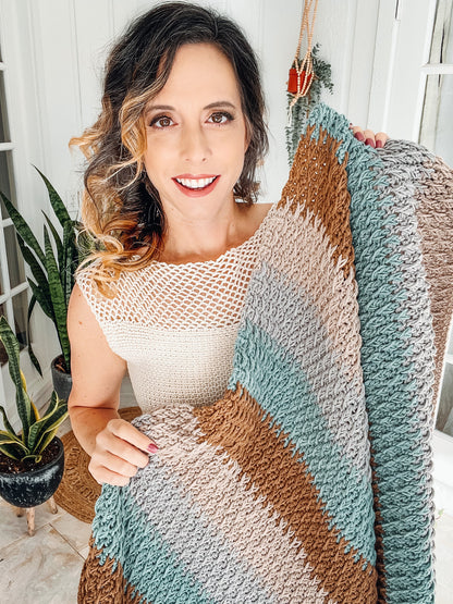Woman in a cream top smiles while holding a textured striped crochet blanket in teal, brown and gray