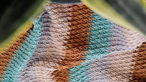 Close-up of textured crochet stitches in brown, teal, gray and beige stripes framed by plant leaves