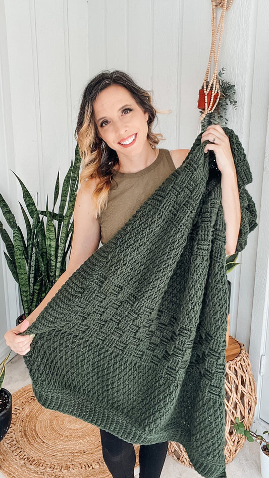 Totally Textured Crochet Blanket pattern: dark green crochet blanket with basketweave texture held up by a smiling woman beside potted snake plants