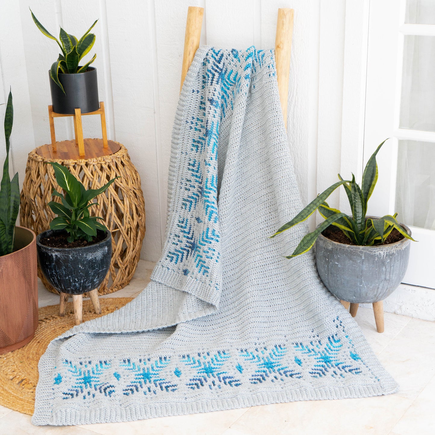A gray crochet blanket with blue patterns is draped over a wooden ladder, surrounded by potted plants on a woven rug, creating an ideal setting for showcasing unique jewellery or presenting as a thoughtful handmade gift.