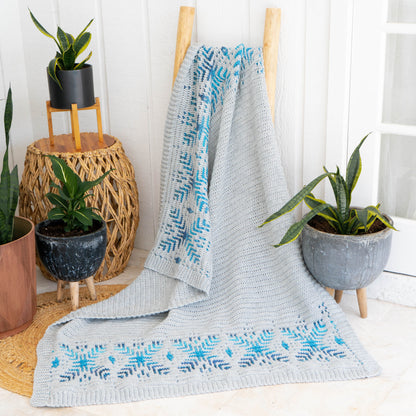 Gray crochet blanket with blue snowflake bands draped over a wooden ladder beside potted plants on a jute rug