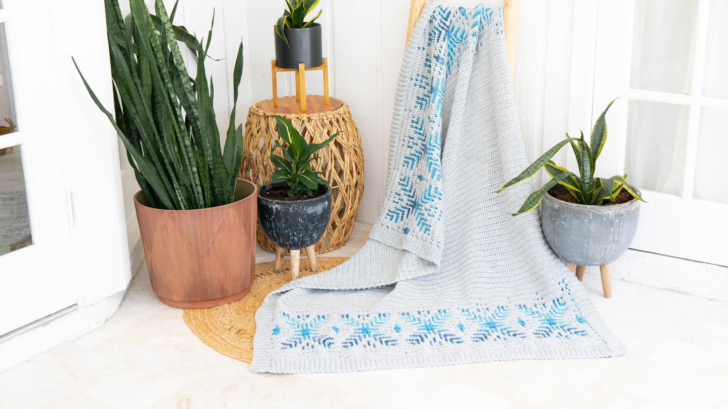 A crochet gray blanket with blue patterns draped over a chair, surrounded by potted plants on a light-colored floor, evokes the charm of a handmade gift.
