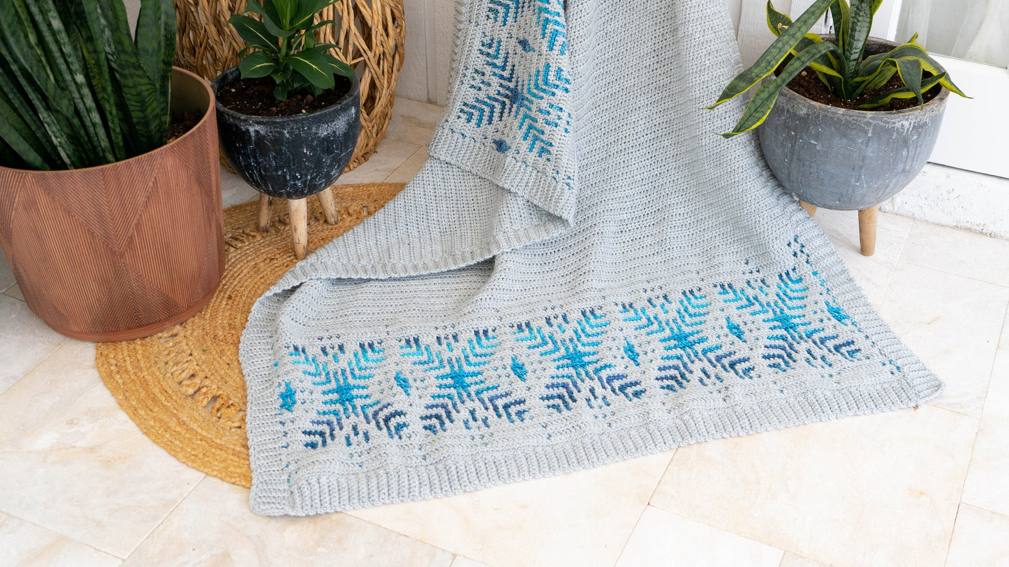 A handmade gift of a gray and blue patterned crocheted blanket is draped between two potted plants on a woven rug on a tiled floor.