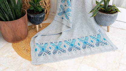 Gray crochet blanket with blue snowflake border spread on a jute rug between potted plants