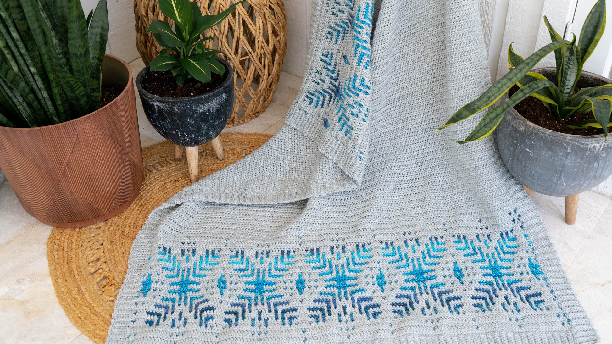 A crocheted blanket with a blue and turquoise geometric pattern draped on a chair, surrounded by potted plants, makes for the perfect handmade gift.
