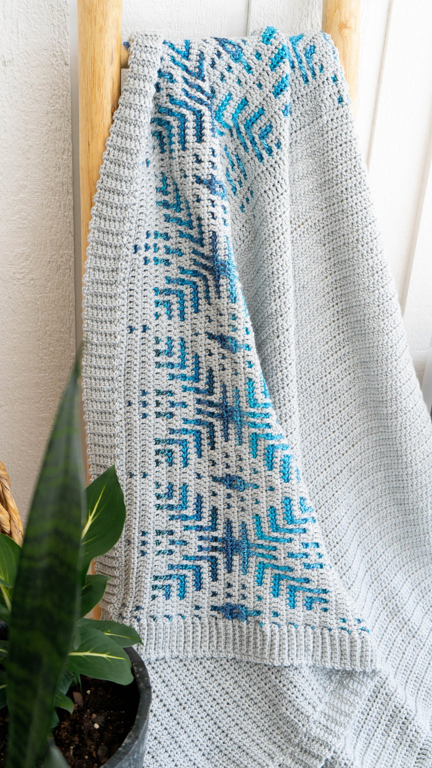 A gray and blue woven blanket with a geometric pattern is draped over a wooden ladder, resembling a cozy handmade gift. A potted green plant is in the foreground.