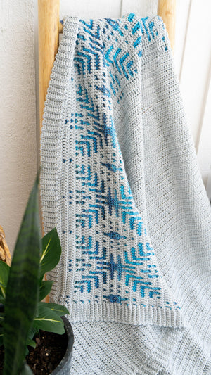 Close-up of gray crochet blanket with blue snowflake motifs and ribbed edge hanging on a wooden ladder