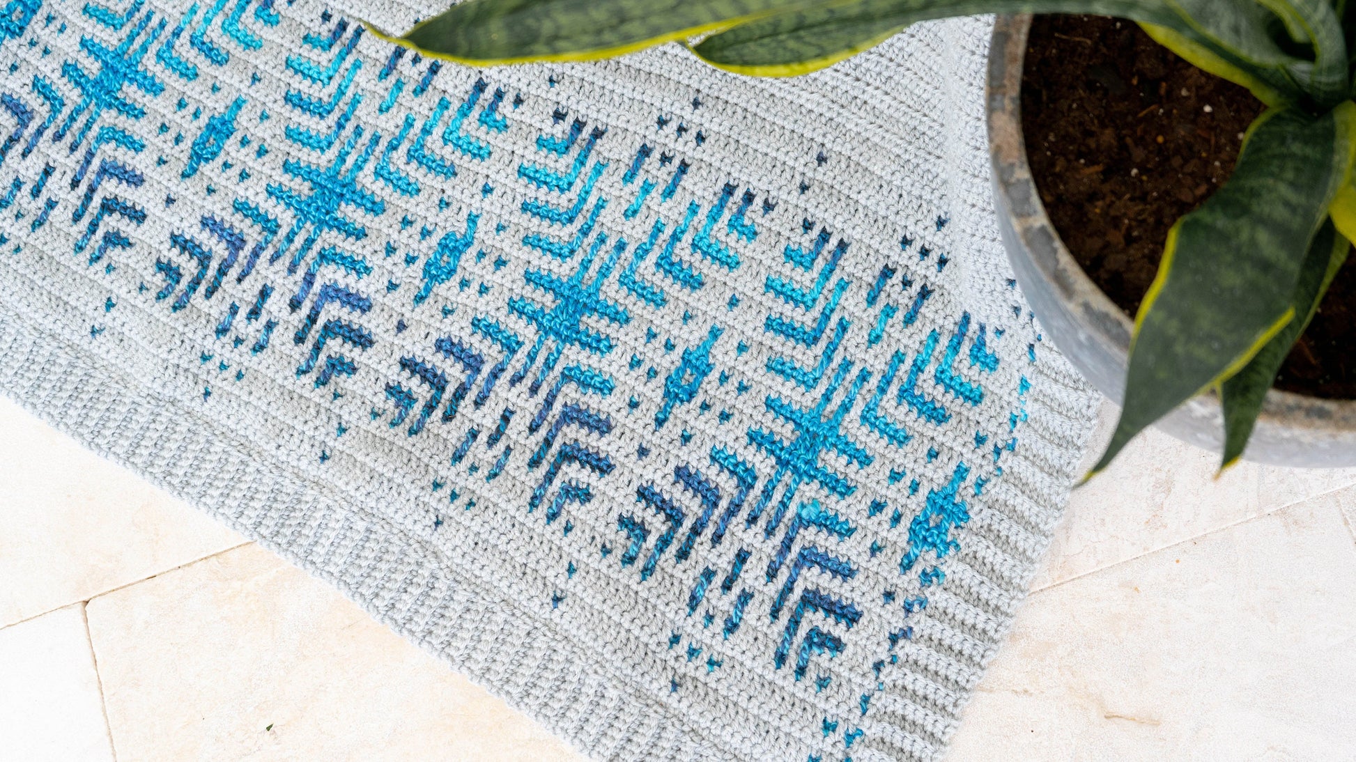 A gray and blue patterned blanket graces the light tiled floor, offering a perfect contrast beside a green potted plant. The scene exudes charm like a piece of unique jewelry in an artisan's handmade gift collection.