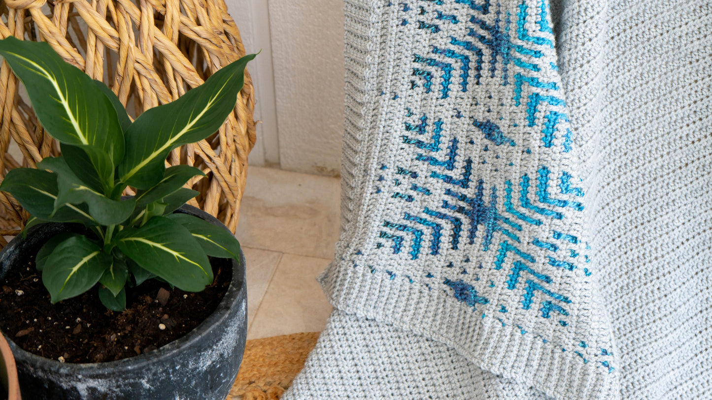 A green potted plant rests beside a blue and grey patterned mosaic crocheted blanket draped next to a wicker chair.