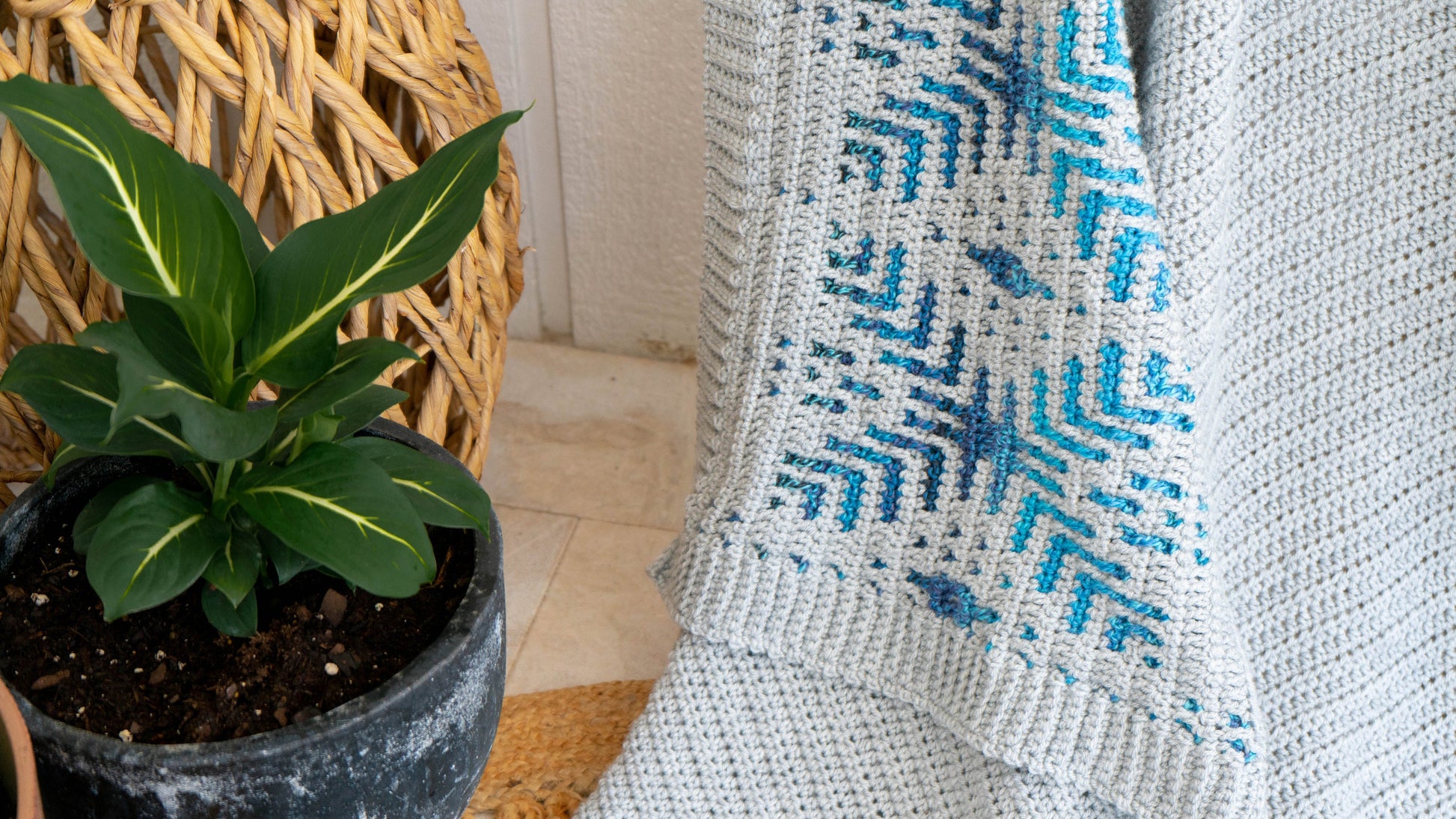 A green potted plant rests beside a blue and grey patterned mosaic crocheted blanket draped next to a wicker chair.
