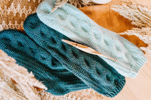 Two teal and one light aqua crochet cabled socks laid flat with a wooden hook and dried pampas grass