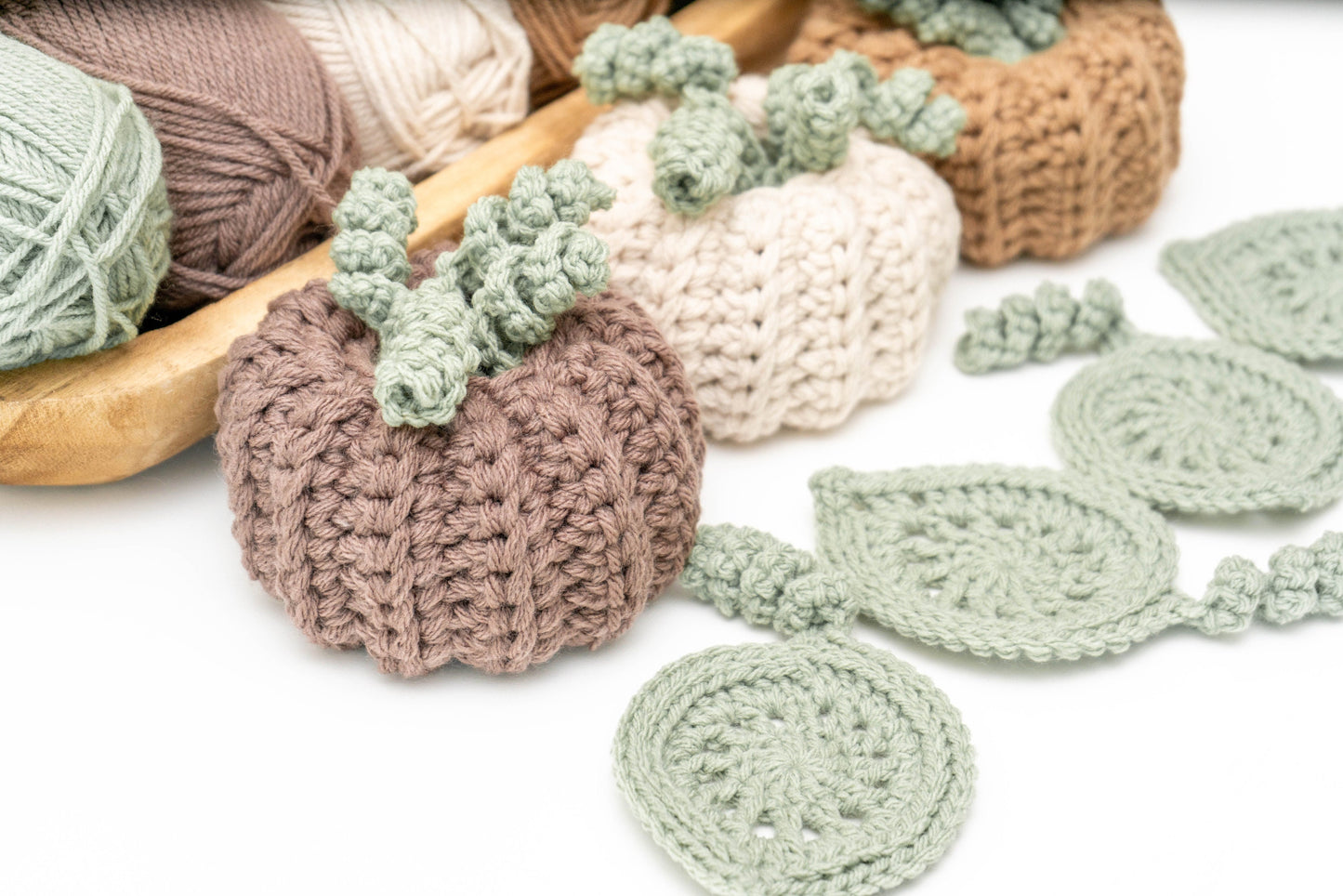 Several skeins of yarn in neutral colors and a crochet project featuring pumpkin and leaf designs, with a crochet hook placed on a white background.