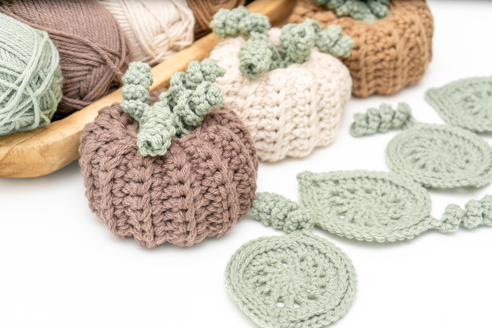 Several skeins of yarn in neutral colors and a crochet project featuring pumpkin and leaf designs, with a crochet hook placed on a white background.