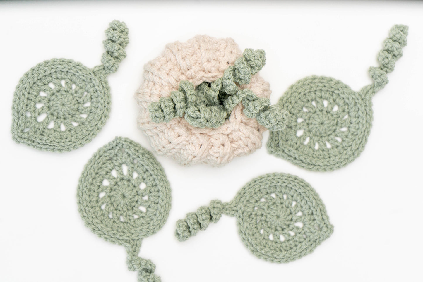 A crocheted beige pumpkin is surrounded by five green crocheted leaves with spiral stems, laid on a white surface—an ideal handmade gift.
