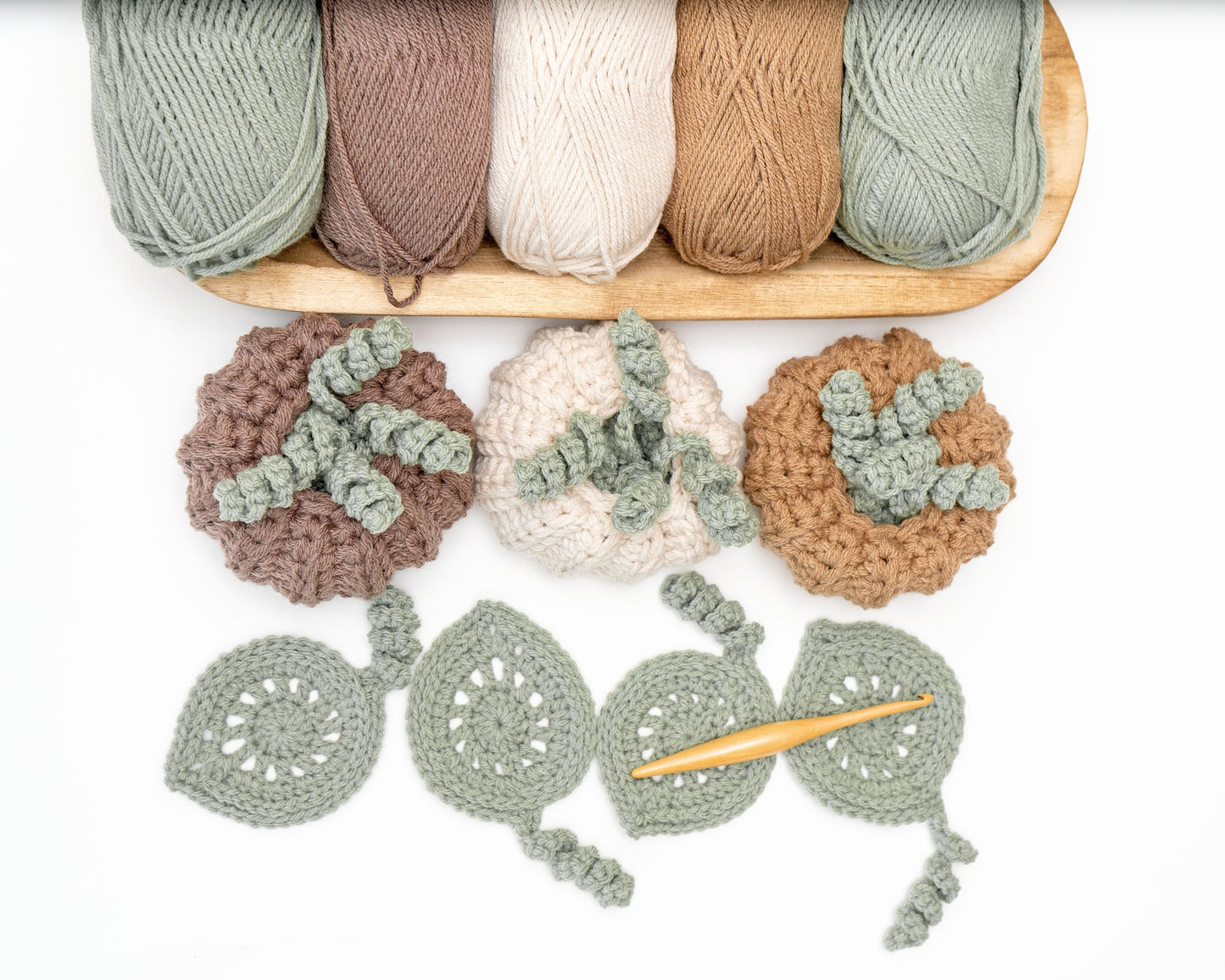 Several skeins of yarn in neutral colors and a crochet project featuring pumpkin and leaf designs, with a crochet hook placed on a white background.
