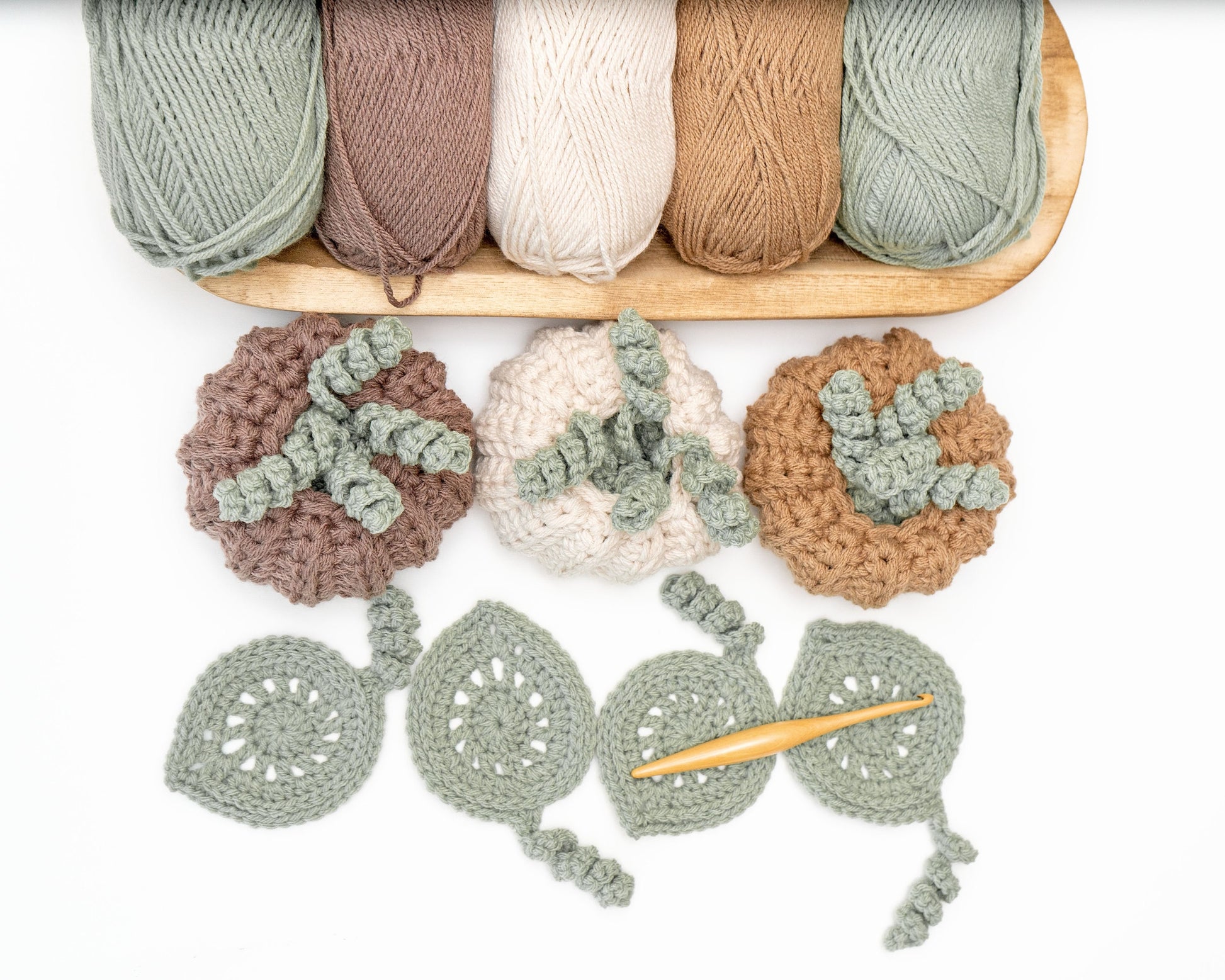 Several skeins of yarn in neutral colors and a crochet project featuring pumpkin and leaf designs, with a crochet hook placed on a white background.