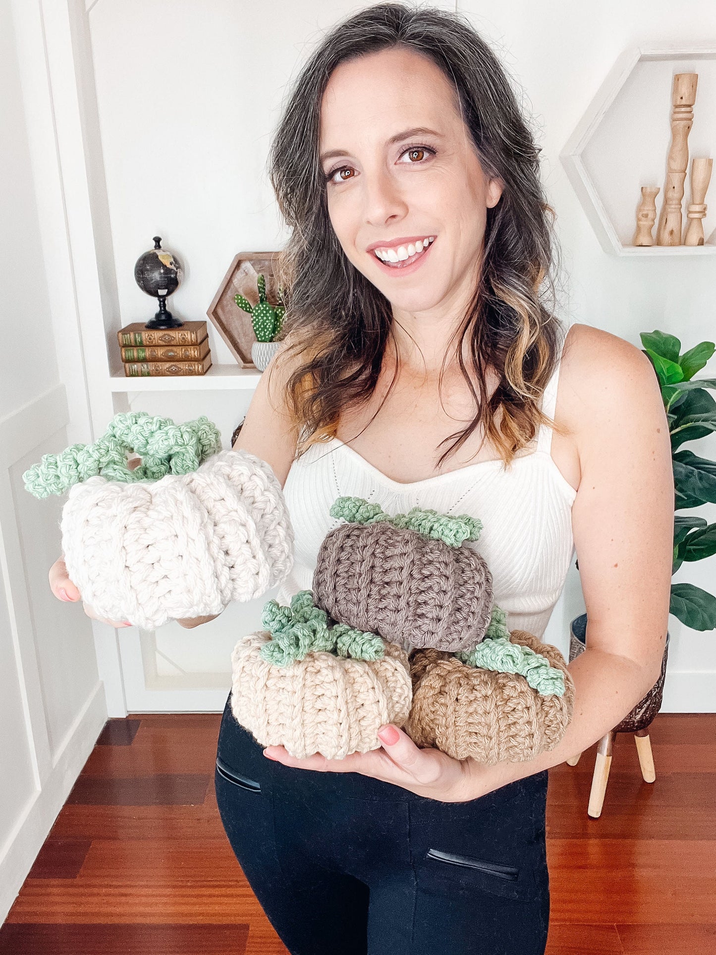 A woman in a white tank top holds four knitted pumpkins in white, brown, and beige. She is standing indoors with a white wall and wooden floor in the background, creating the perfect setting to showcase these handmade gifts.