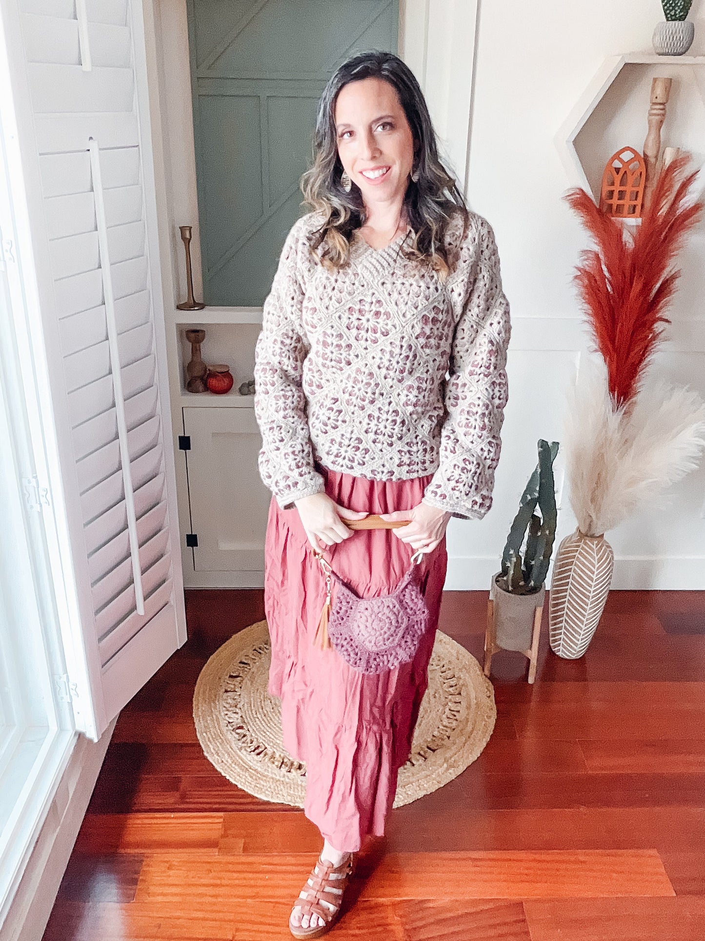A woman in a patterned sweater and long red skirt stands indoors, smiling with a round purse. She exudes charm, accessorized with unique jewellery. Decor includes a geometric shelf, pampas grass, and vases on the wooden floor.