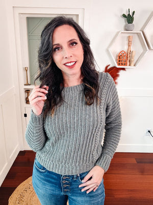 Gray green ribbed crochet sweater worn by a woman in jeans standing in a white room with shelves