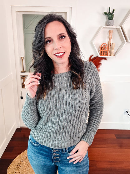 Gray green ribbed crochet sweater worn by a woman in jeans standing in a white room with shelves