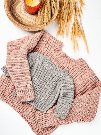 Gray crochet child sweater laid on a larger pink one with wheat stalks and a candle on white
