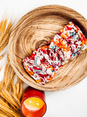 Chunky multicolor crochet ear warmer in red, orange and teal in a wooden bowl beside wheat and a candle