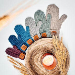 Crochet mittens in burgundy, blues, grays, and beige fanned around a wooden bowl with a candle and wheat
