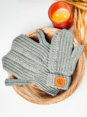 Gray crochet mittens with cable and ribbed texture in a wooden bowl next to a lit candle and wheat